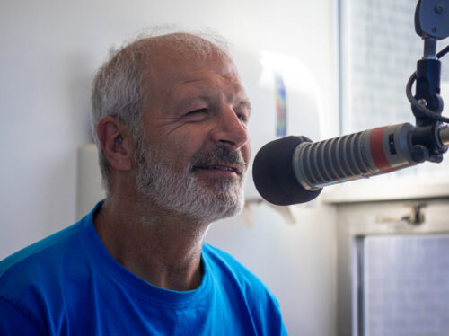 Lucien Comeau, une homme grisonnant en chandelle bleu parlant au micro. Il est dans un studio de radio.