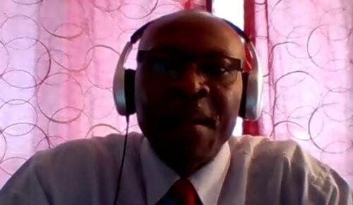 Crispin Mbaya, un homme noir, vêtu dune chemise blanche, cravate rouge et veston gris, posant devant un rideau rose, lors d'une rencontre de presse en Zoom.