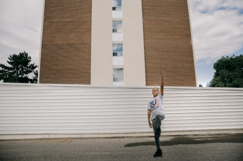 Young Lay-Up participant posing outside, near an apartment building. Their arm and leg are raised.