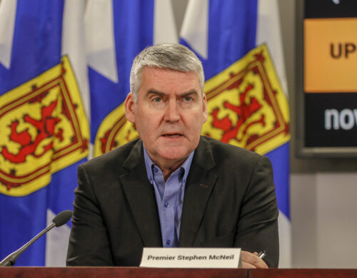 Premier Stephen McNeil sits at a wooden desk in front of a microphone at a press conference with Nova Scotia flags lined up behind him.