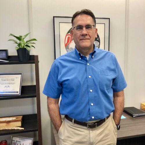 Bill Pavich stands in his office, wearing a blue button up shirt, khakis, and glasses. 