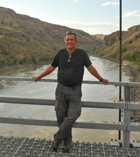 Russ Dawson stands on a bridge crossing a river, wearing a t-shirt and khakis.