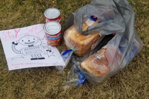 Photo of typical grocery delivery with two loaves of bread and two cans of food. Delivery also includes a handwritten note by a child that reads, "stay saff'