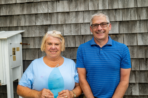 Beverly Hugli et Paul Boucher posent et sourient. Le fond est fait de tuiles en bois typique des maisons acadiennes. 