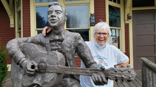A smiling woman has her arm around a statue of a man playing guitar.