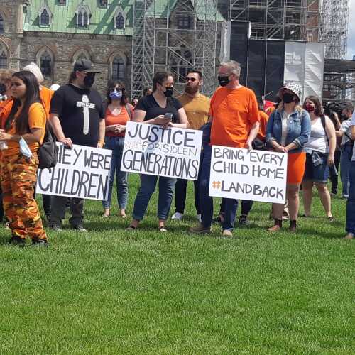 A crowd of people on the lawn on Parliament hill holding signs of protest in orange shirts. 