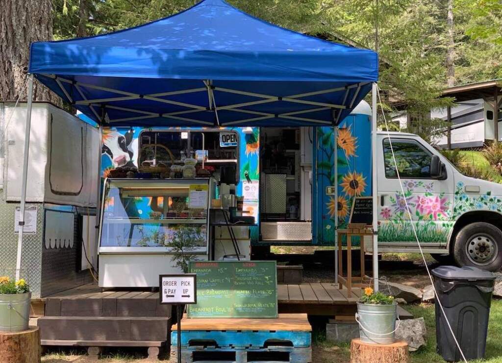 There are steps leading up to platform, covered by a blue canopy. A food counter and til are inside. A vividly painted truck with Sunflowers painted on the side, is behind the platform and off to one side is a trailer/kitchen