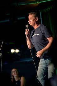 Humoriste vêtu d'un T-Shirt bleu et de Jeans faisant de la comédie Stand-up sur estrade.
