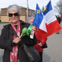 Dame aux cheveux gris, lunettes à soleil, manteau noir et foulard rouge avec un petit tambourin vert et des drapeaux acadiens.