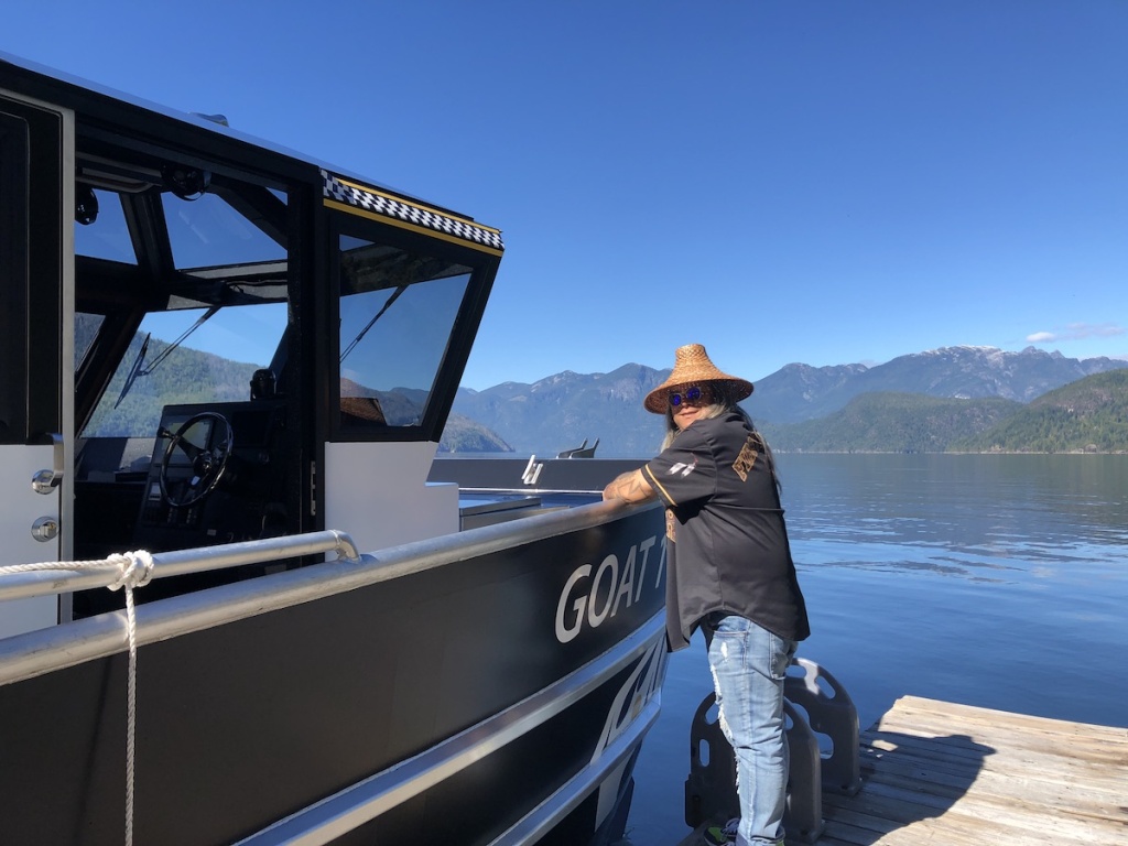 A man in an Indigenous woven cedar hat leans on a docked boat on a sunny day.