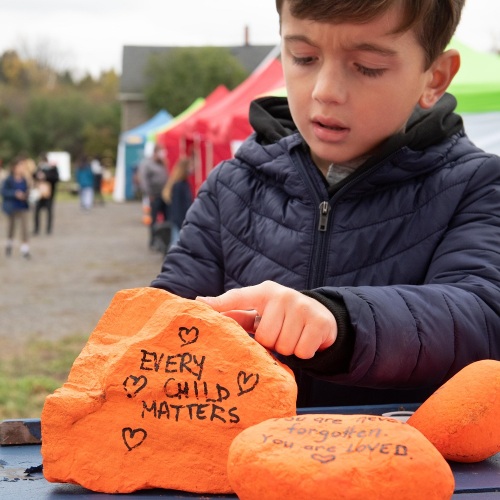 A young child with brown hair wearing a dark blue jacket is seen touching a rock which is painted orange and has the slogan "Every Child Matters" written in black.