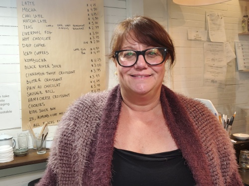 A woman wearing glasses, stands in front of a menu board in a cafe