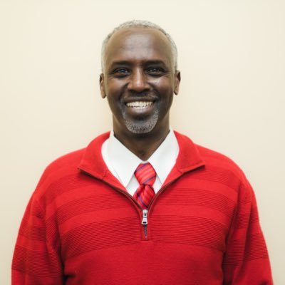 Ali Duale un homme noir souriant avec les cheveux grisonnants portant un pull rouge une chemise blanche et une cravate rouge. Il poser devant un fond neutre.