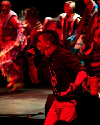 A man wearing a black sweatshirt and light coloured pants is seen singing into a microphone. In the background, traditional Indigenous performers are seen dancing.