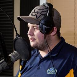 Homme avec casquette, chandail bleu et écouteurs parlant dans un micro de radio.