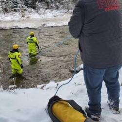 Une homme sur le rivage et deux hommes dans une rivière avec des équipements de sauvetage.