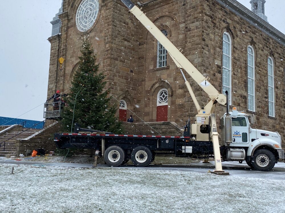 Des gens installent un arbre de Noël géant avec l'aide d'un gros camion en avant d'une église en roches.