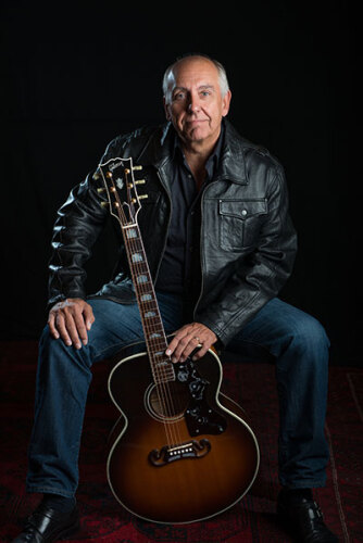 Homme avec gilet en cuir, jeans et chemise bleu assis avec une guitare.