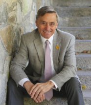 homme aux cheveux grisonnant assis sur des marches en pierre, il porte une chemise blanche, cravate rose et verste de costume gris-beige