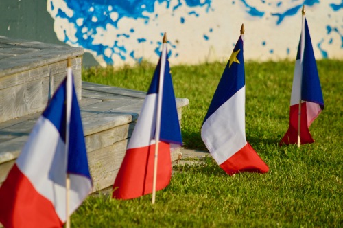 Quatre petits drapeaux acadiens plantés par terre en avant d'un estrade en bois.