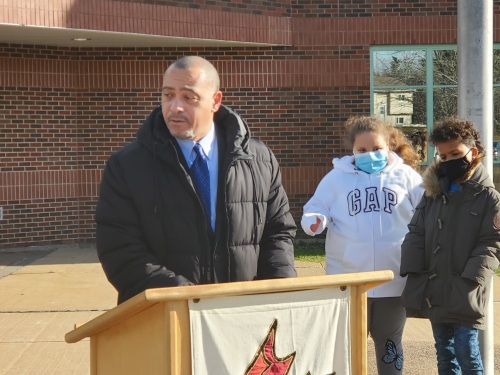 A man speaks at a podium outside a school as students watch in front of a brick building.