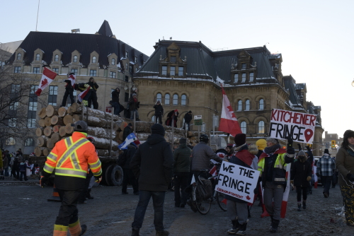 A lumber truck is seen with protestors atop, waving Canada flags. A group of protestors marches on foot beside them, holding signs reading, "We the Fringe."