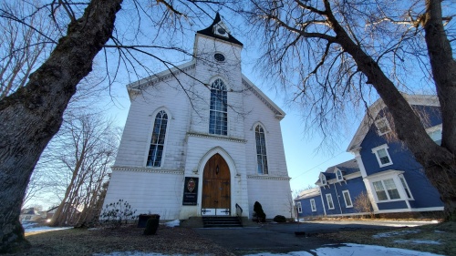 The exterior of a white church on a sunny day. There is a row of blue buildings next to the church.