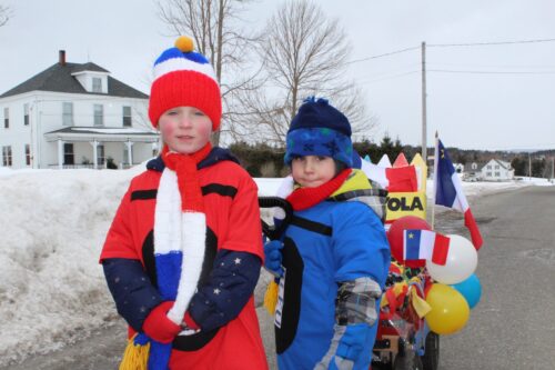 Deux enfants habillés aux couleurs acadiennes tirant un chariot décoré dans la rue en hiver.