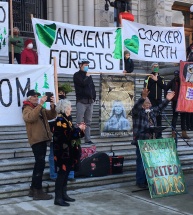 homme âgé devant un pupitre et micro les mains levées, arrirère plan des gens avec de banderoles avec des inscriptions en faveur des forêts anciennes et de la planète