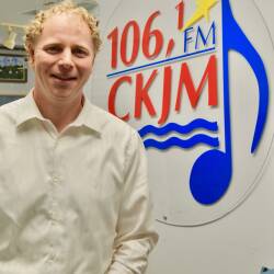 Un homme portant une chemise blanche en avant du logo de Radio CKJM attaché à un mur.
