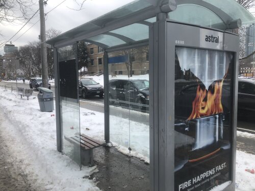 A bus shelter with clear glass and grey sides on a bed of white snow on the side of a main road with cars parked.