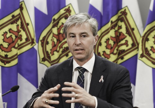 A man speaks at a desk in front of a row of Nova Scotia flags