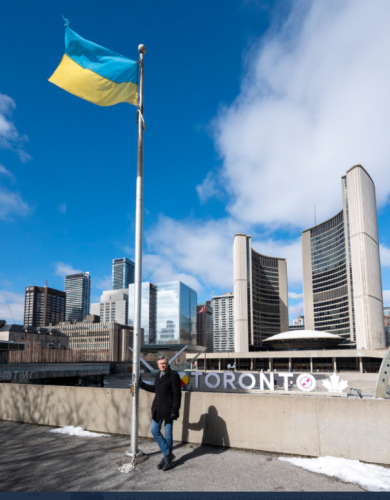 A man in a black coat and jeans standing on the right side of a large pole with a yellow and blue flag on top.