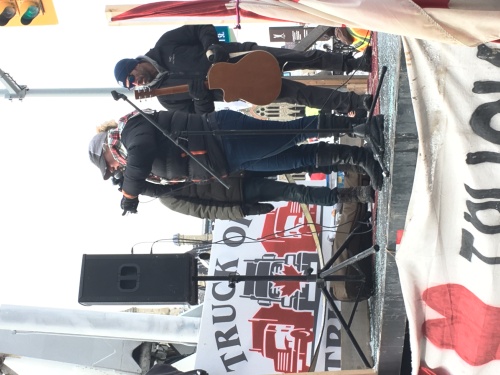 A woman is seen speaking into a microphone, standing on a stage made from a truck bed, surrounded by pro-convoy signage, with two performers standing at her side.