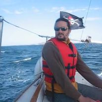 Un homme avec lunettes à soleil et portant un gilet de sauvetage rouge sur un voilier en pleine mer.