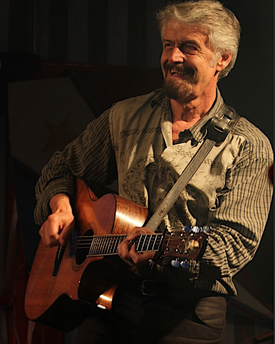 Homme aux cheveux gris avec chemise et culottes brunes jouant de la guitare.