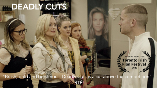 Four women and a man all dressed in white, gold and black stand looking at each other with a poster of a woman with blonde hair in the background wall.