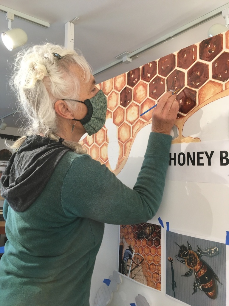 A woman with white hair and a green sweater is painting honeycombs on a panel.
