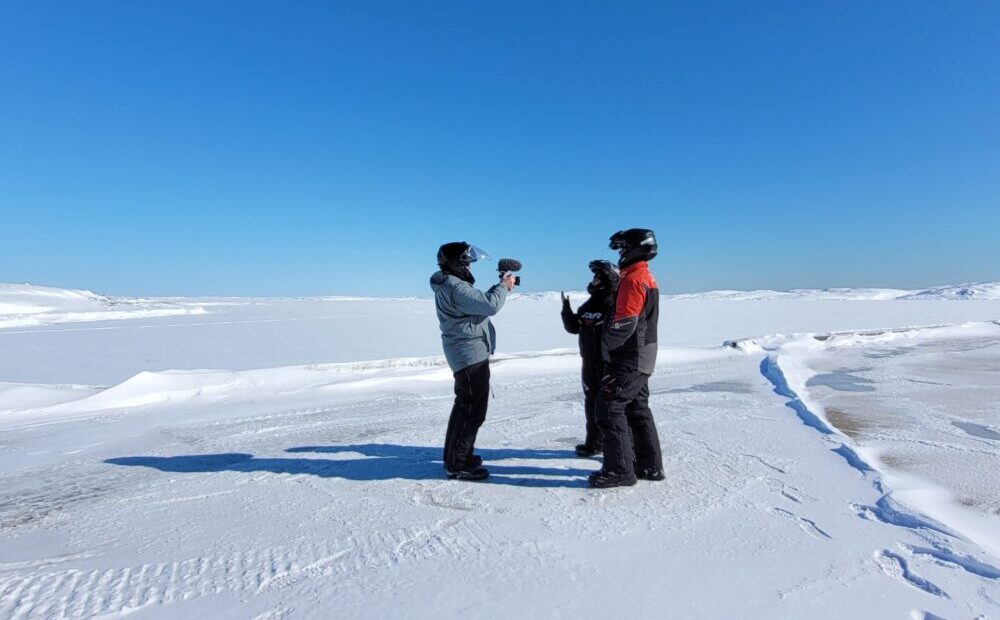 Des plaines de neige, un ciel bleu et deux personnes en habit de motoneige se font filmer par une autre personne habillé de la même manière.