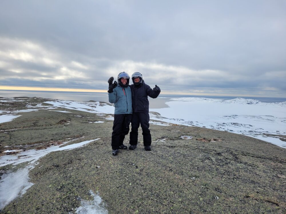 Deux hommes portant des casques de motoneige sur un morne dégarni de neige. L'horizon à perte de vue avec le fleuve au loin.