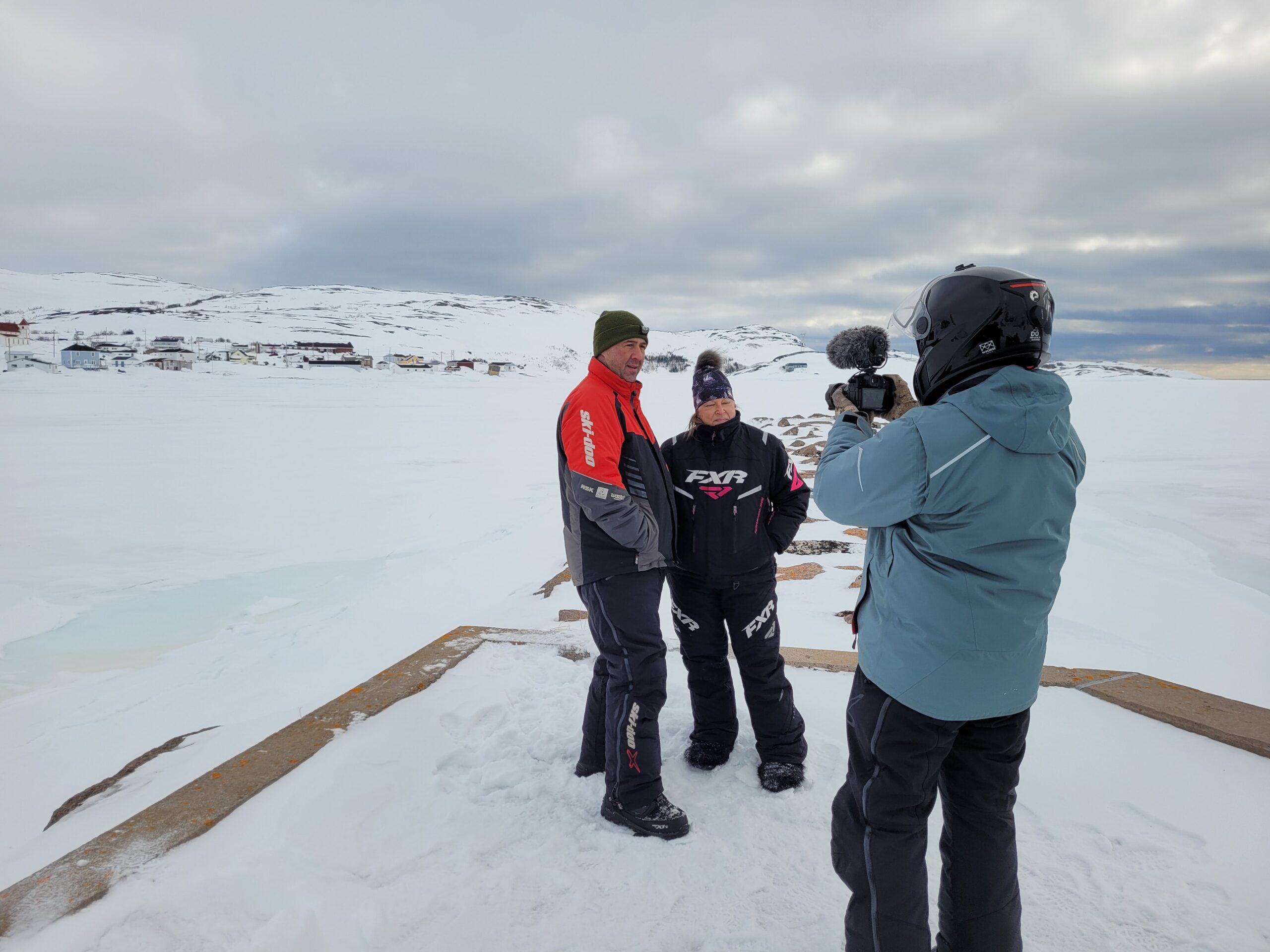 Deux personnes en habit de motoneige se font filmer devant un décor blanc de neige.