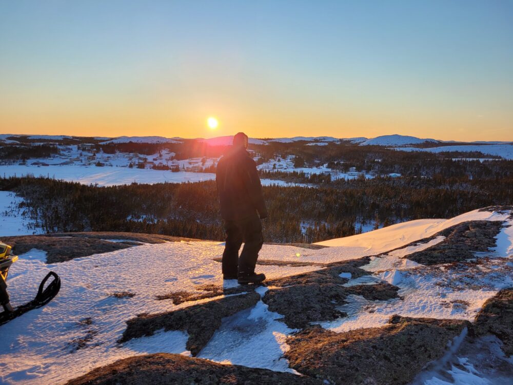 Un homme à contre-jour regarde au loin un coucher de soleil orangé au sommet d'un morne rocheux parsemé de neige.