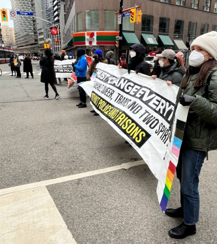 People standing at a road intersection holding up a white banner with a grey building containing a colourful store of green and orange sits in the background.