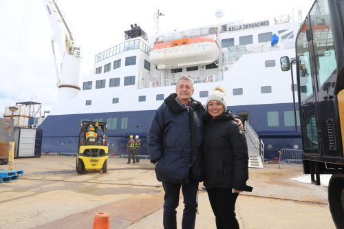 Un couple habillé de manteaux d'hiver pose plein pied devant un navire en train d'être déchargé.