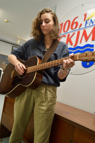 Une jeune femme aux cheveux longs portant une chemise et des pantalons verts jouant de la guitare en avant du logo de Radio CKJM.