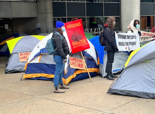 Three people stand in front of multicoloured pitched tents outside of a building entrance. Two people on the right are holding a white sign with black text.