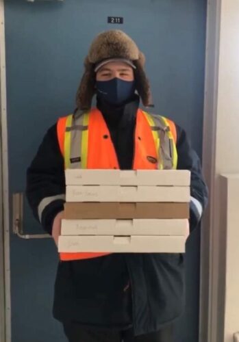 Un élève habillé chaudement pour l'hiver tient cinq boîtes de pizza devant une porte menant vers l'extérieur.