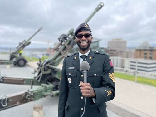 Sgt. Ainsley Francis is standing in front of the canon for a photo smiling in his official uniform, with a microphone in his hand as this was taken during our interview.