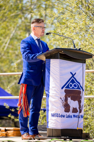 A man stands at a podium at Willow Lake Métis Nation on a sunny day with trees behind him.