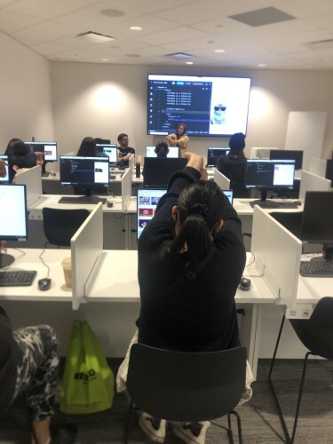 A row of students in a classroom take a stretch break in front of computers. There is a large screen at the front of the room.